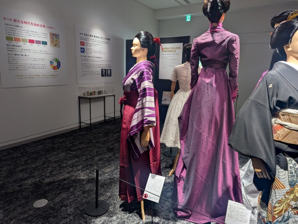 Mannequins in vibrant purple and pink dresses on display in a gallery, with informational panels on the wall behind them.